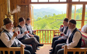 چند مرد گیلک شاد با کلاههای نمدی در حال نوشیدن چای لاهیجان در قهوهخانهای سنتی رو به باغات چای